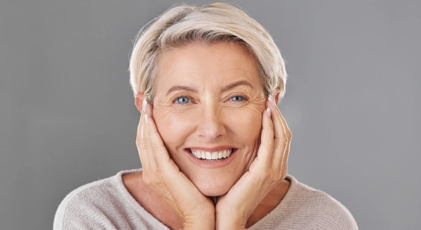 Smiling mature woman with white hair touching her face against gray background, dental wellness portrait.